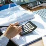 A person working on financial calculations using a calculator and laptop at an office desk.